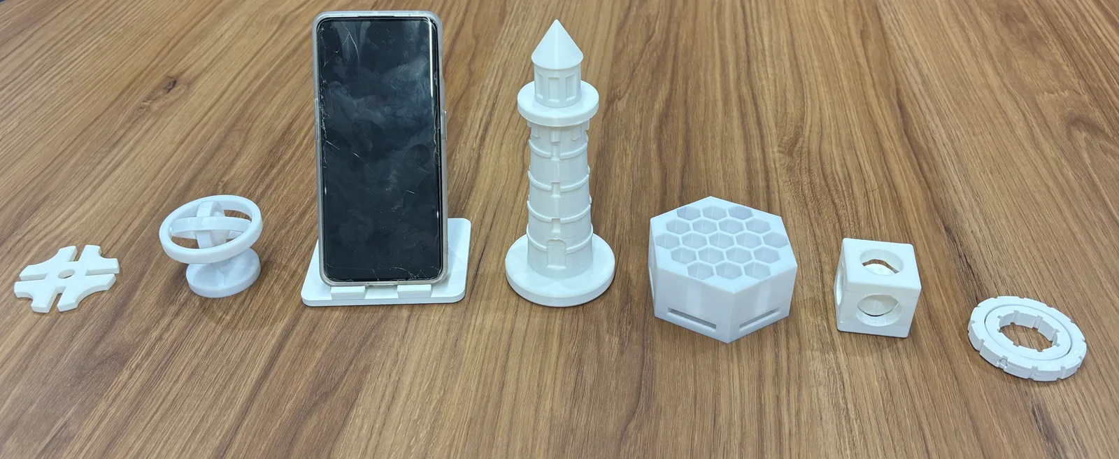 A row of white 3D-printed parts generated by MAC, including a phone stand, lighthouse, honeycomb organizer, gyroscope and ball-in-cage